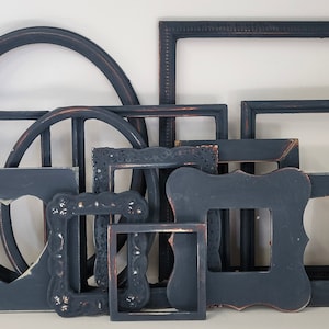 May include: A collection of various dark gray picture frames in different shapes and sizes. Some frames have ornate details, while others are simple. One frame has a heart-shaped cutout. The frames are arranged against a white background.