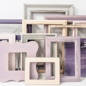 May include: A collection of empty picture frames in various sizes and styles, painted in shades of white, lavender, and purple. Some frames have distressed finishes, and one features a wire mesh backing. The frames are arranged against a white background.