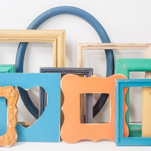 May include: A collection of colorful, empty picture frames in various shapes and sizes. The frames are painted in shades of blue, green, orange, and cream, with some showing distressed finishes. A blue, arched frame sits in the background.