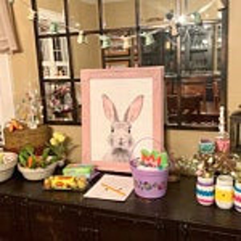 Peut inclure: Une impression encadr&eacute;e d'un lapin rose, un seau violet avec des accents floraux et des &oelig;ufs de P&acirc;ques d&eacute;coratifs sont pos&eacute;s sur une surface en bois sombre. Un panier de fleurs et d'autres d&eacute;corations de P&acirc;ques sont &eacute;galement visibles.