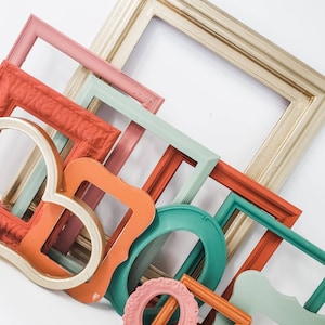 May include: A collection of colorful picture frames in various shapes and sizes. The frames are painted in shades of coral, teal, pink, gold, and orange. Some frames have ornate details, while others are simple. The frames are arranged on a white surface.