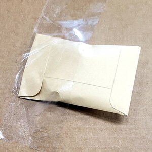 May include: A small, light brown paper envelope is partially encased in a clear plastic bag. The envelope is sealed and has a rectangular shape with rounded corners. The background is a light brown surface.