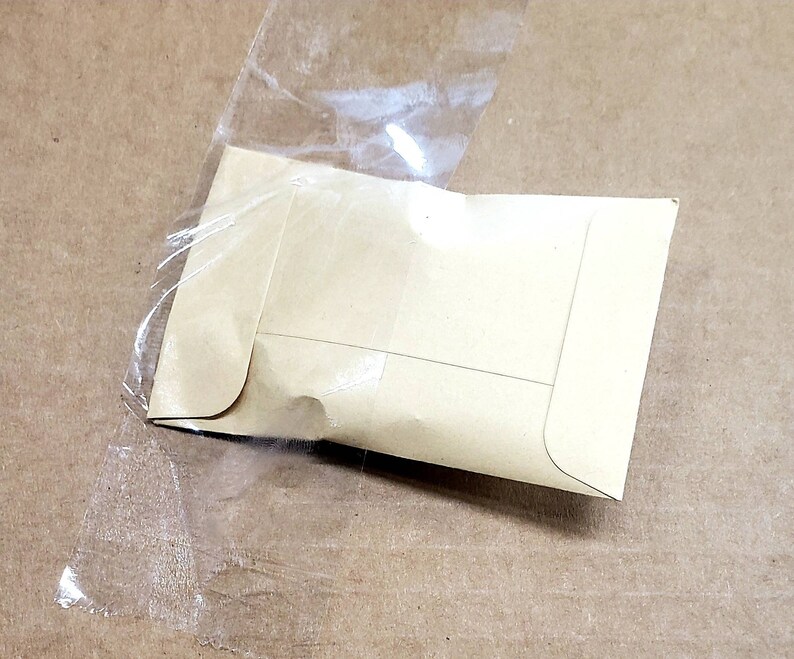 May include: A small, light brown paper envelope is partially encased in a clear plastic bag. The envelope is sealed and has a rectangular shape with rounded corners. The background is a light brown surface.