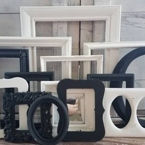 May include: A collection of various picture frames in black and white. The frames are in different shapes and sizes, including rectangular, oval, and circular. Some frames have ornate details, while others are simple. The frames are arranged against a light-colored wooden background.