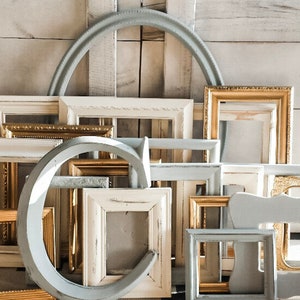May include: A collection of various picture frames in different shapes and sizes. The frames are in shades of white, gold, and gray, with some having ornate details. The background is a light-colored wooden wall.