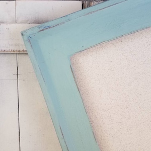 May include: A corner of a rectangular picture frame with a distressed, light blue painted finish. The frame has a thick, beveled edge and a neutral-colored backing. The frame is positioned at an angle against a white wooden surface.