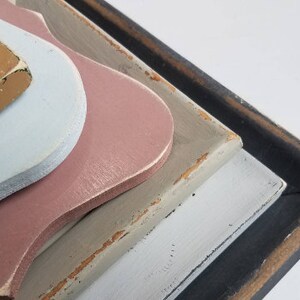 May include: Close-up of layered, decorative wooden shapes in various colors. The shapes include light blue, pink, tan, and gray, with a black frame. The edges show a distressed, aged appearance.