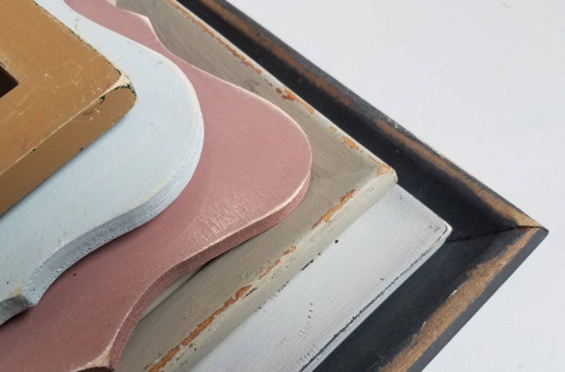 May include: Close-up of layered, decorative wooden shapes in various colors. The shapes include light blue, pink, tan, and gray, with a black frame. The edges show a distressed, aged appearance.