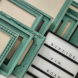May include: A collection of teal picture frames with various ornate and beaded designs. A black and white letter board spells out "RIPTIDE PIN BOARD". The frames are arranged at different angles, showcasing their details.