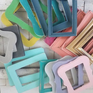 May include: A collection of colorful, vintage-style picture frames in various shapes and sizes. The frames are painted in shades of green, blue, pink, gold, and gray, with a distressed finish. The frames are arranged on a white wooden surface.