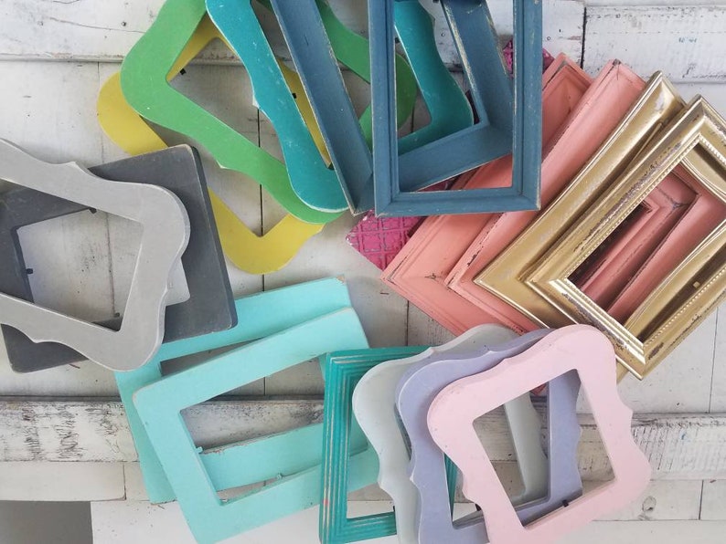 May include: A collection of colorful, vintage-style picture frames in various shapes and sizes. The frames are painted in shades of green, blue, pink, gold, and gray, with a distressed finish. The frames are arranged on a white wooden surface.