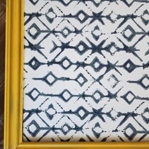 May include: A framed artwork featuring a repeating geometric pattern in shades of blue and white. The artwork is encased in a distressed yellow wooden frame, adding a touch of rustic charm. The design is a series of diamonds and lines.