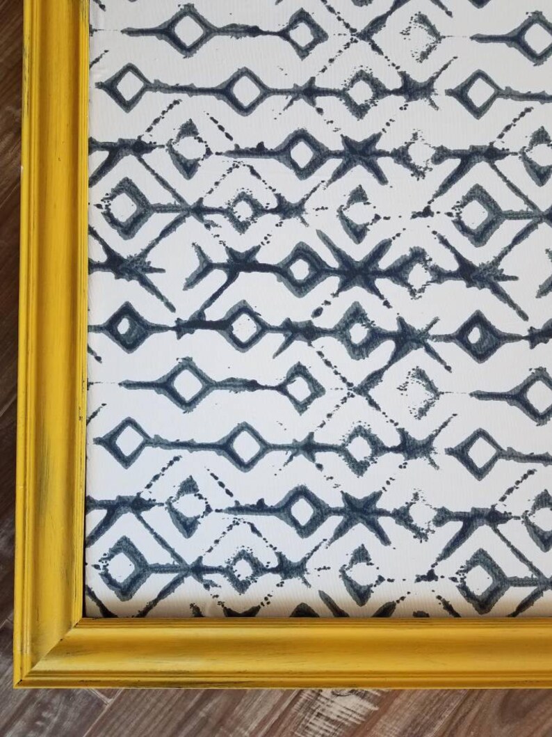 May include: A framed artwork featuring a repeating geometric pattern in shades of blue and white. The artwork is encased in a distressed yellow wooden frame, adding a touch of rustic charm. The design is a series of diamonds and lines.