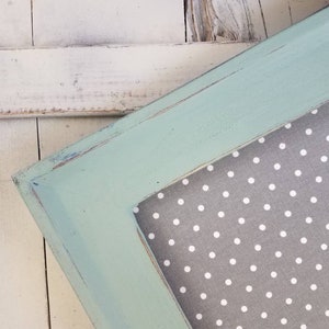 May include: A square picture frame with a distressed, light blue painted finish. The frame surrounds a gray fabric with white polka dots. The frame is set against a white, weathered wooden background.
