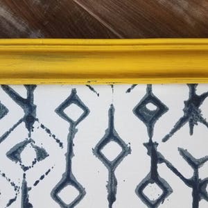 May include: A framed artwork featuring a white background with a repeating pattern of dark blue geometric shapes. The frame is a bright yellow with a distressed finish, adding a pop of color and texture to the piece. The artwork is likely a print or painting.