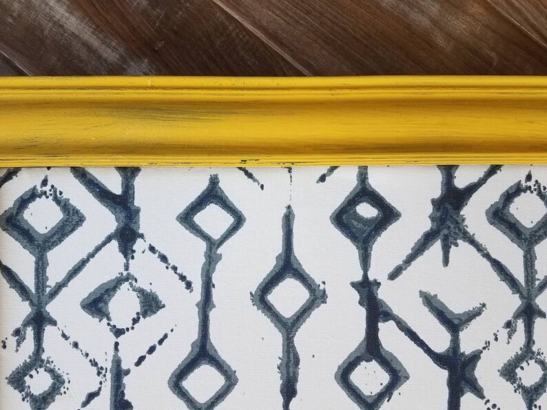 May include: A framed artwork featuring a white background with a repeating pattern of dark blue geometric shapes. The frame is a bright yellow with a distressed finish, adding a pop of color and texture to the piece. The artwork is likely a print or painting.