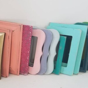 May include: A collection of colorful picture frames in various shapes and sizes. The frames are painted in shades of gray, gold, pink, teal, blue, green, and yellow. Some frames have ornate designs, while others are simple.