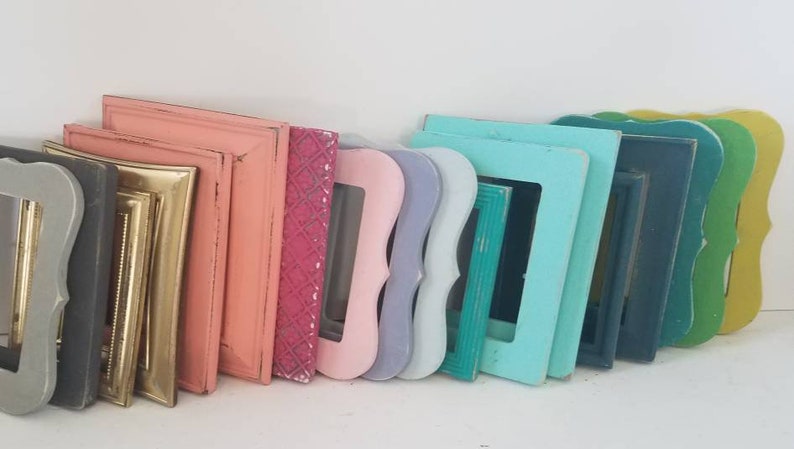 May include: A collection of colorful picture frames in various shapes and sizes. The frames are painted in shades of gray, gold, pink, teal, blue, green, and yellow. Some frames have ornate designs, while others are simple.