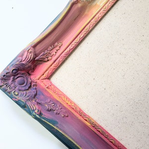 Peut inclure: Un coin d'un cadre photo d&eacute;coratif et orn&eacute;. Le cadre est peint avec un d&eacute;grad&eacute; de couleurs, notamment du rose, du violet, du bleu et de l'or. Le cadre pr&eacute;sente des motifs floraux et des volutes d&eacute;taill&eacute;s. Le fond est une toile.