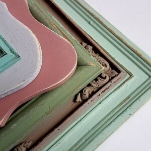 May include: A stack of decorative wooden frames in various colors. The frames include a distressed teal, green, pink, and gray. Some frames have ornate corner details, and all have a vintage, weathered appearance.