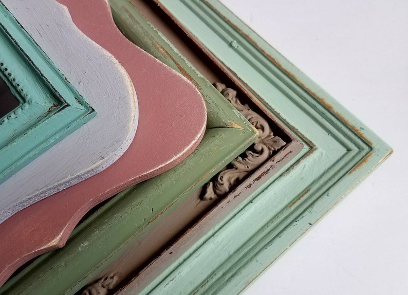 May include: A stack of decorative wooden frames in various colors. The frames include a distressed teal, green, pink, and gray. Some frames have ornate corner details, and all have a vintage, weathered appearance.