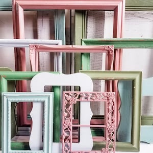 May include: A collection of vintage picture frames in various sizes and colors. The frames are painted in shades of pink, green, blue, and white, with some featuring ornate details. The arrangement suggests a focus on home decor and wall art.