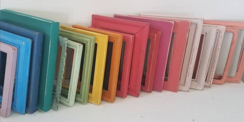 May include: A collection of colorful picture frames in various sizes and shades, including blue, green, yellow, orange, pink, and white. The frames are leaning against a white surface, showcasing a range of vibrant colors and styles.