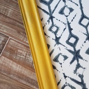 May include: A golden yellow picture frame corner with a decorative edge, positioned diagonally against a patterned background. The background features a white surface with a repeating dark blue geometric design, and a wood-grain surface.