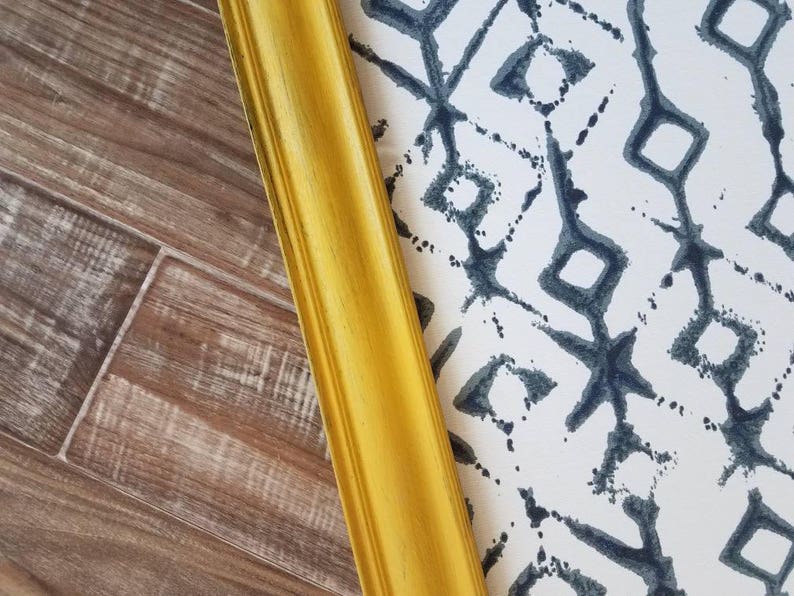 May include: A golden yellow picture frame corner with a decorative edge, positioned diagonally against a patterned background. The background features a white surface with a repeating dark blue geometric design, and a wood-grain surface.