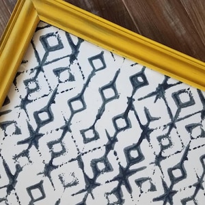 May include: A framed print with a yellow painted wooden frame. The print features a repeating pattern of dark blue diamond shapes on a white background. The frame has a distressed finish, adding a vintage feel to the artwork.