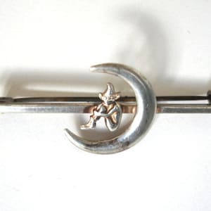May include: Silver-toned brooch depicting a crescent moon with a seated figure. The figure, wearing a pointed hat, is seated within the moon's curve. The brooch has a pin closure on the reverse.