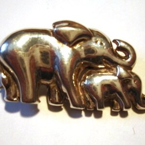 May include: A silver-coloured brooch depicting a family of elephants. The brooch shows a large elephant followed by a smaller one, with a baby elephant in front. The elephants have detailed trunks and ears.