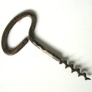 May include: A vintage metal corkscrew with a spiral worm and a looped handle. The corkscrew is a tarnished silver colour and rests on a white surface. The handle has text around the loop.