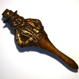May include: A vintage brass bottle stopper in the shape of a man wearing a hat and coat. The figure is detailed with facial features and clothing textures. The stopper has a long, tapered handle.
