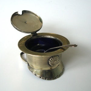 May include: An antique silver-toned salt cellar with a hinged lid and a small spoon. The salt cellar has a textured design and a dark blue interior. The lid is slightly ajar, revealing the contents. The design is reminiscent of a top hat.