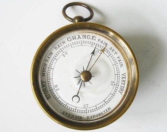 May include: A vintage aneroid barometer with a brass casing and a white face. The face features a needle pointing to "Fair" and text indicating weather conditions like "Stormy" and "Very Dry". A small ring is at the top for hanging.