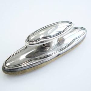 May include: A silver shoe shine brush featuring an oval handle and a rectangular base. The brush has a light brown, textured pad for polishing shoes. The handle is raised and has a decorative, curved design.