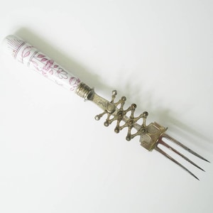 Könnte beinhalten: Eine antike Gabel aus Silber und Porzellan mit floralem Design. Der Griff ist weiß mit rosa Blumenmustern. Die Gabel hat drei Zinken und einen dekorativen Metallmechanismus. Ein einzigartiges Vintage-Küchenutensil.