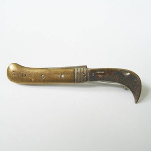 May include: A vintage brass folding knife with a curved blade. The handle is a warm gold color with three small rivets. The blade is partially open, revealing a dark, curved cutting edge. The knife appears to be well-worn.