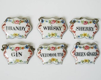 May include: Six vintage white ceramic liquor labels with floral designs and metal loops. Each label features a different liquor name in black text: Brandy, Whisky, Sherry, Gin, Vermouth, and Green Ginger.