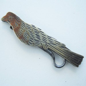 May include: A vintage metal bird-shaped letter opener. The opener features a detailed design with a silver body and a bronze-colored head. The bird has a curved tail and a hook-shaped handle.