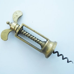 May include: A vintage brass corkscrew with a spiral worm and butterfly-shaped levers. The corkscrew has a rectangular frame and a decorative base. The metal has a warm, aged patina, suggesting it is an antique.