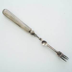 Peut inclure: Un ustensile de service argenté avec un manche cannelé, une petite cuillère et une fourchette à deux dents. L'ustensile est probablement vintage et conçu pour un aliment spécifique, comme les olives ou les cornichons.