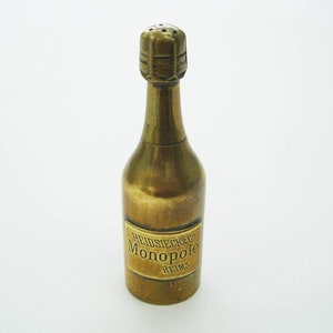 May include: A brass-colored salt or pepper shaker shaped like a champagne bottle. The bottle has a label that reads "Heidsieck & Co Monopole Reims". The top of the bottle has small holes for dispensing.