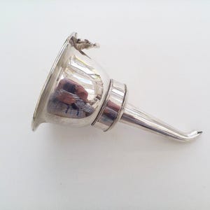 Puede incluir: Un embudo de metal de color plateado pulido con una abertura superior ancha y acampanada y un pico cónico y curvado. La superficie reflectante del embudo muestra un reflejo distorsionado del entorno y se observa un pequeño detalle decorativo en el borde.