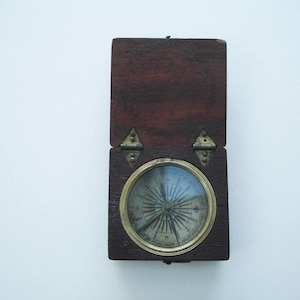 May include: An open, antique compass in a dark wood case. The compass face is visible through a glass cover, with a gold-colored metal ring. The case is rectangular with brass hinges and a clasp.