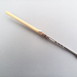 May include: A silver and cream cocktail fork with a three-pronged end. The handle is cream-coloured, leading to a silver band with decorative detailing. The fork is designed for serving appetizers or garnishes.