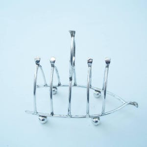 Könnte beinhalten: Ein silberfarbener Toastständer mit einem einzigartigen Design. Er verfügt über vertikale Stäbe, die von einer gebogenen Basis und kleinen kugelförmigen Füßen getragen werden. Der Ständer besteht aus glänzendem Metall und ist für mehrere Toastscheiben konzipiert.