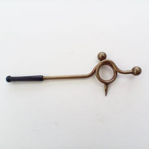 May include: A brass-colored metal tool featuring a central circular ring, two small spherical finials, and a downward-pointing metal spike. The handle is a long, thin metal rod finished with a smooth, dark black cylindrical tip at the end.