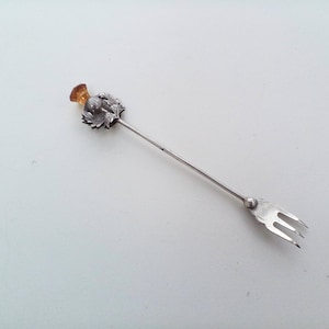Peut inclure: Une fourchette à cocktail argentée dotée d'un long manche fin surmonté d'un motif décoratif en métal en forme de chardon et d'une petite pierre translucide de couleur ambre à l'extrémité, terminée par une tête à trois dents.